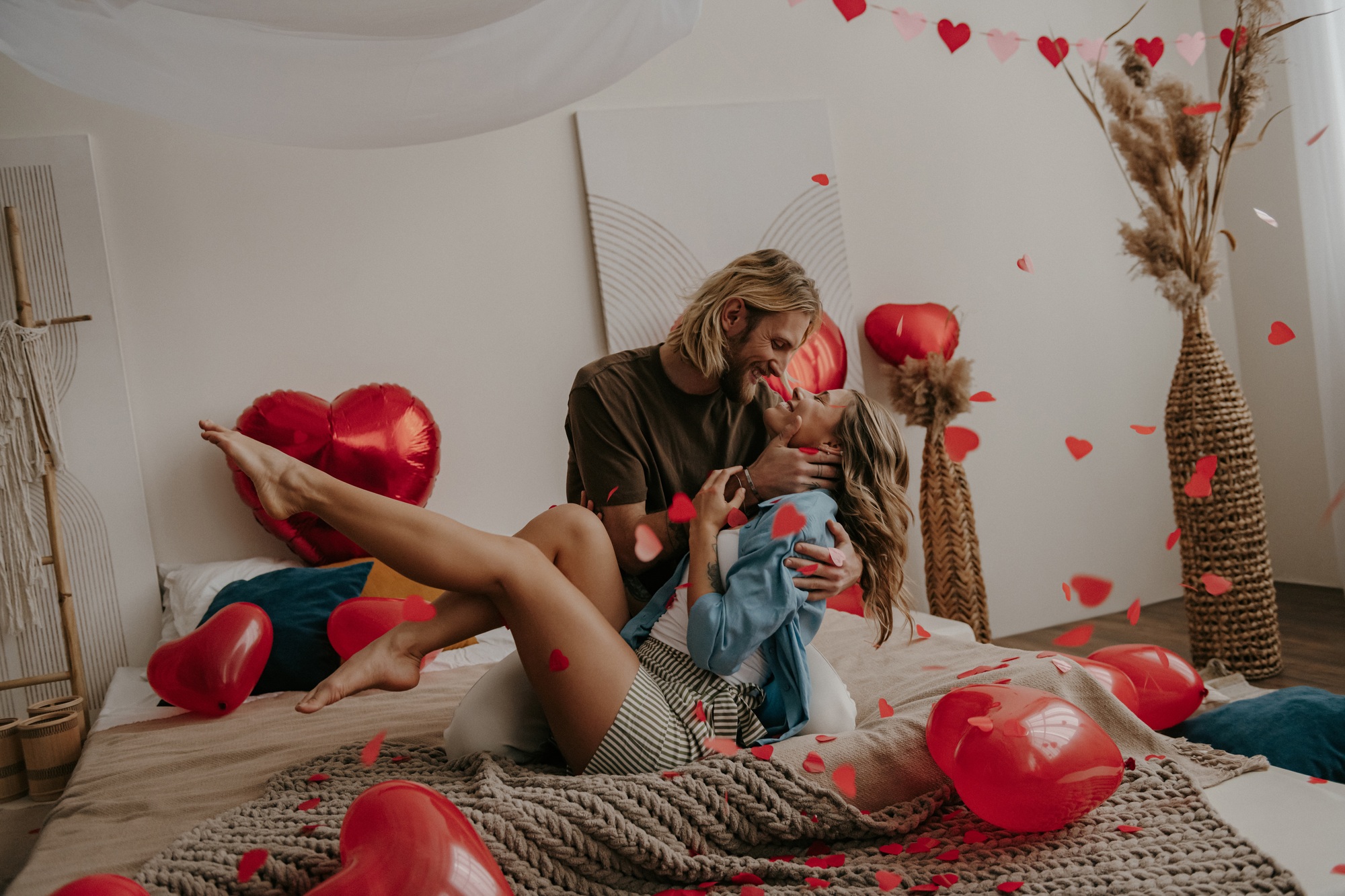 Romantic loving couple sitting on bed and kissing while red heart shape confetti flying around
