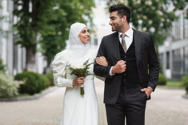 happy muslim bride in wedding hijab and white dress holding bouquet near groom outside