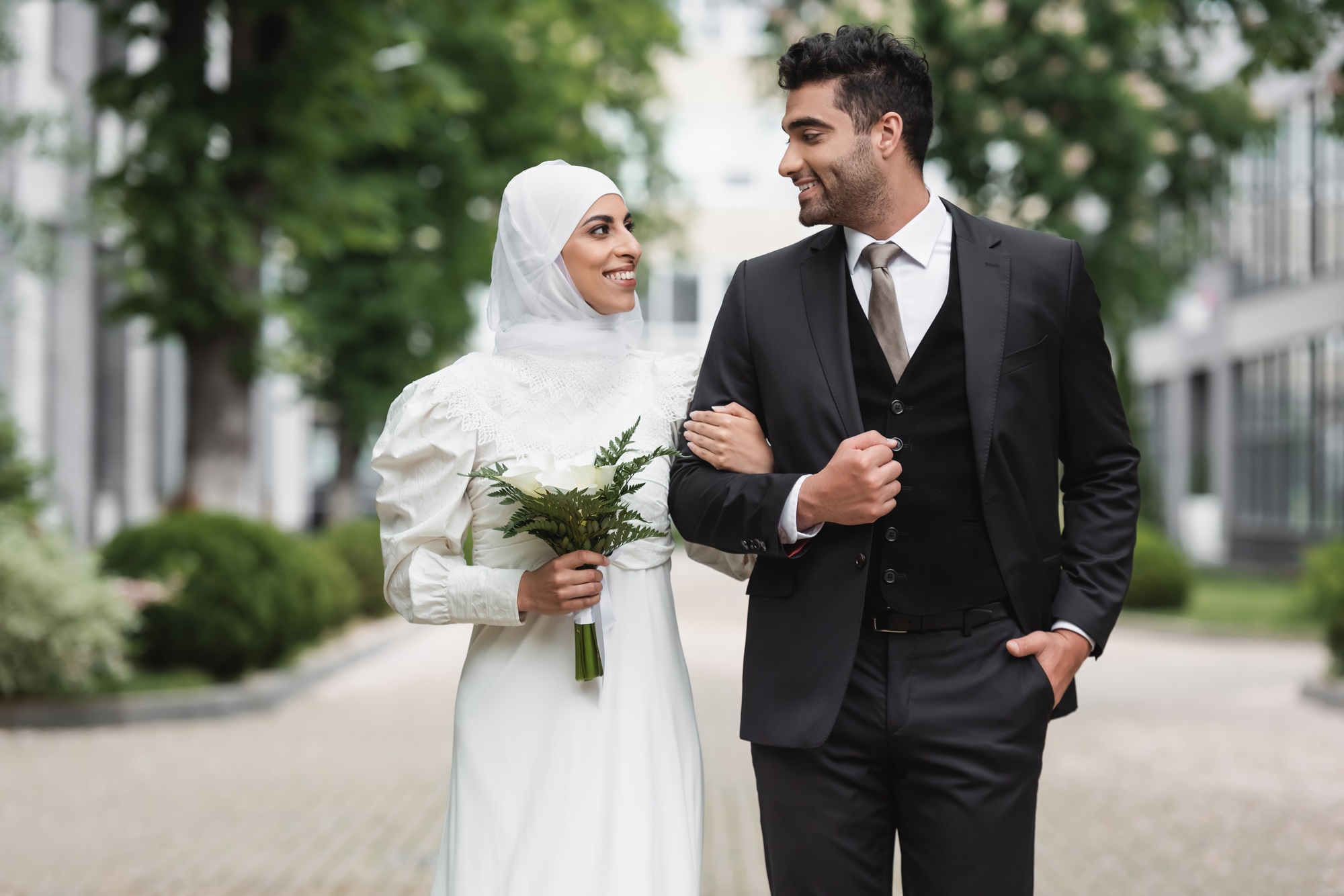 happy muslim bride in wedding hijab and white dress holding bouquet near groom outside