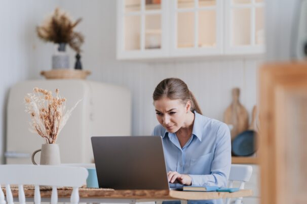 Focused blonde caucasian woman in shirt sits at desk with laptop looks at screen in disbelief